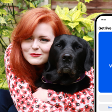 Lucy is crouching down and hugging a black Labrador in front of a garden wall. Next to her and the dog is a large iPhone screen showing the homepage of the Be My Eyes app, which is photoshopped into the picture. 