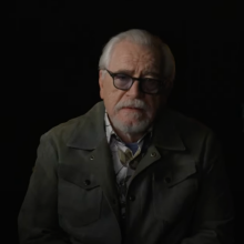 Actor Brian Cox on a mostly dark set, wearing glasses and looking serious.