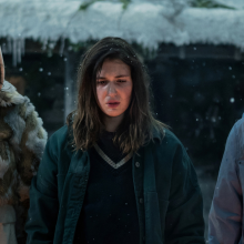 Three girls wearing thick coats stand in front of a fire while it snows. 
