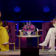 A woman wearing a yellow suit interviews a woman wearing a white suit. 