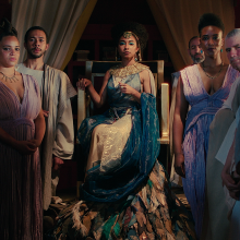 An Africa queen sits on her throne surrounded by her subjects, wearing Ancient Egyptian robes. 