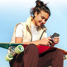 Woman sitting down with longboard on lap checking her phone.
