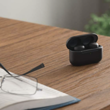 Black Amazon Echo Buds on a brown wood desk with a pair of glasses on top of a book on bottom left, tablet on upper left side
