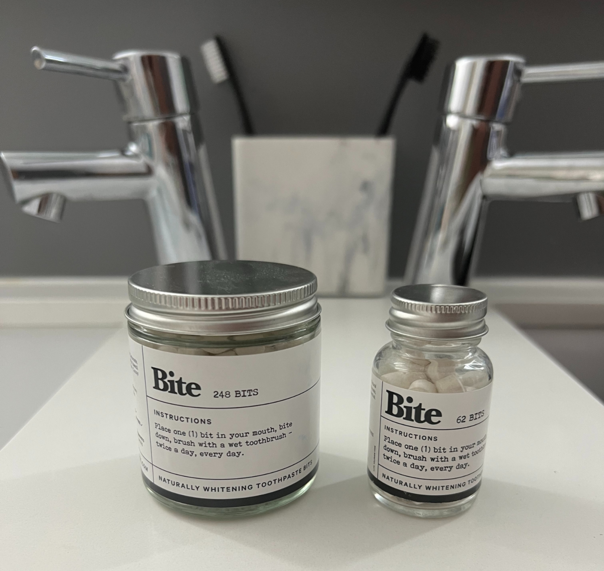 Two sizes of Bite Toothpaste Bits jars on bathroom counter with faucets and toothbrushes in background