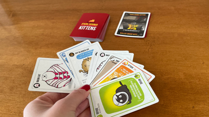 Manicured hand holding a bunch of exploding kittens cards next to the game box